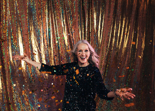 Happy Senior Woman In Black Dress Standing Under Confetti Against A Golden Backdrop