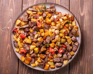 Large plate with fried potatoes with mushrooms and meat on a wooden background, top view. Homemade food for the whole family.