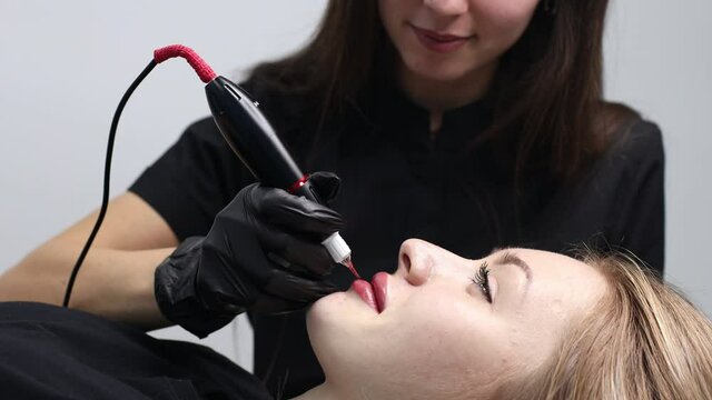 Cosmetologist applying pink pigment permanent tattoo on female lips with tatooing needle machine at salon