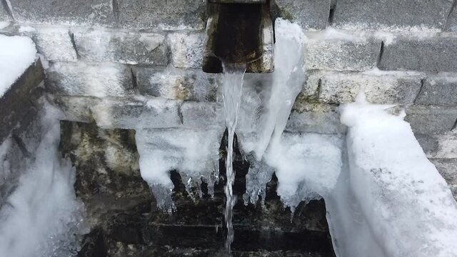 Steadicam shot of winter gushing spring out of a brick wall by granite gutter with ice and icicles around