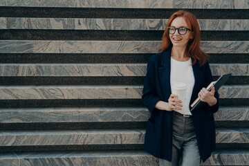 Woman dressed formally drinks takeaway coffee holds digital tablet and notepad