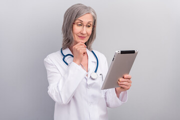 Photo of positive thoughtful mature lady doctor wear white coat spectacles smiling reading modern gadget isolated grey color background