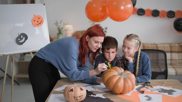 charming woman with a girl and a boy have fun drawing funny pumpkin faces on the table with drawings for a festive autumn party