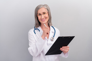 Photo of cute positive senior woman doc dressed white uniform glasses signing clipboard smiling isolated grey color background