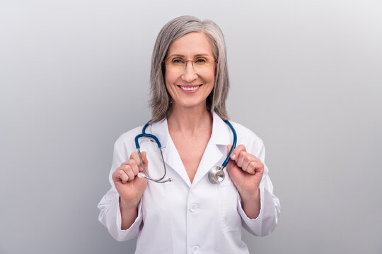 Photo of positive happy mature lady doctor wear white coat spectacles smiling arms phonendoscope isolated grey color background