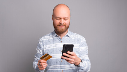 Cheerful bearded man in casual making payment with credit card and smartphone.