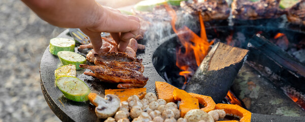 Close-up detail view of chef flipping by tongs tasty crispy marinated quails and vegetables grilled at round steel iron firepit hearth table surface brazier with burning firewoods. Barbecue yard home - obrazy, fototapety, plakaty