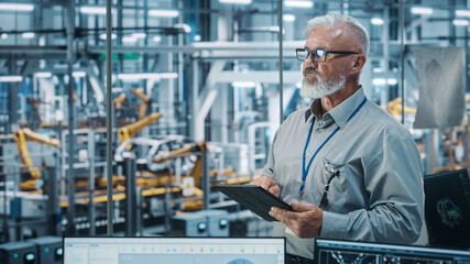 Car Factory Office: Portrait of Senior White Male Chief Engineer Using Tablet Computer in Automated Robot Arm Assembly Line Manufacturing High-Tech Electric Vehicles. Medium Shot