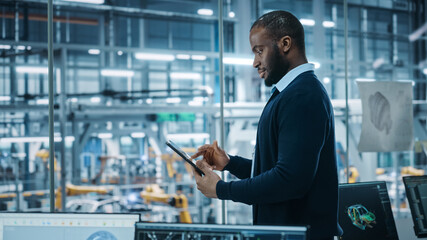 Car Factory Office: Portrait of Successful Black Male Chief Engineer Using Tablet Computer in Automated Robot Arm Assembly Line Manufacturing High-Tech Electric Vehicles. Medium Side View