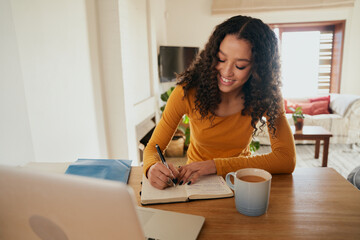 Multi-cultural female happily writing in notebook. Young professional remote working with laptop in modern apartment