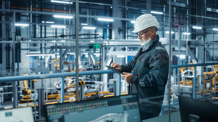 Car Factory: Male Automotive Engineer Wearing Hard Hat, Standing Using Tablet Computer. Monitoring,...