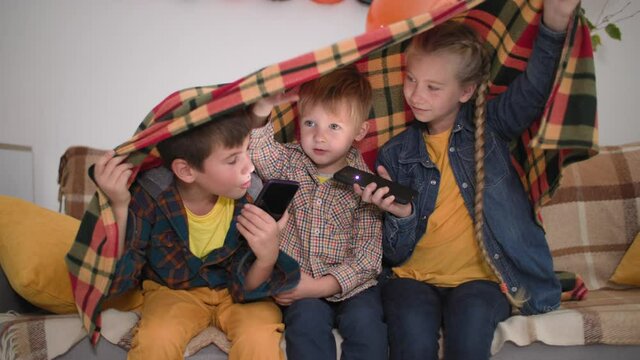 fun pastime for children,cheerful boys and girl with phones in their hands shine on their faces and tell scary stories covered with blanketlaugh and look at camera, while sitting on couch in party