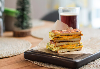Roasted sandwich cheese and scrambled eggs on paper with coffee glass cup on wooden board. Dark background.
