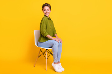 Photo of positive lovely lady sit chair posing wear green shirt jeans footwear isolated yellow color background