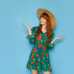 Red haired girl in green summer dress and straw hat is posing with hands raised.