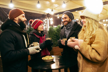 Happy smiling friends with cups of mulled wine having fun, spending time together at winter fair at...