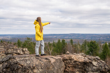 A tourist in a yellow jacket and gray pants stands on the top of a mountain in a valley of the mountains and points into the distance.