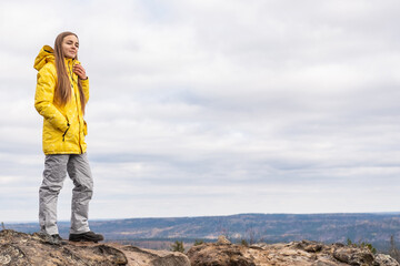 A smiling tourist, in a yellow jacket and gray pants, stands on the top of a mountain in a valley of mountains and admires the beauty of nature.