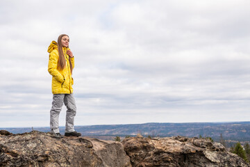A smiling tourist, in a yellow jacket and gray pants, stands on the top of a mountain in a valley of mountains and admires the beauty of nature.