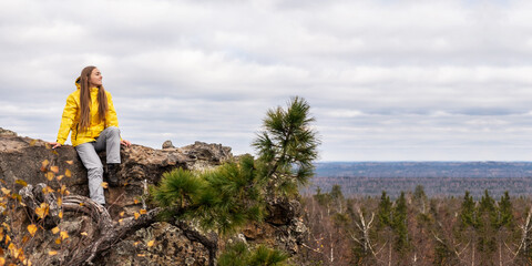 A smiling tourist, in a yellow jacket and gray pants, sits on the top of a mountain and admires the beauty of nature. Panoramic image.