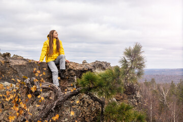 Fototapeta premium A smiling tourist, in a yellow jacket and gray pants, sits on the top of a mountain, in a beautiful valley with mountains and cloudy skies, admiring the beauty of nature.