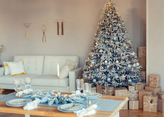 Bright living room decorated with New Year decor. Christmas tree with gold and silver decorations and lots of wrapped gift boxes under it. Christmas table setting in foreground
