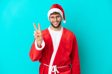 Fototapeta na wymiar Young man disguised as Santa Claus isolated on blue background smiling and showing victory sign
