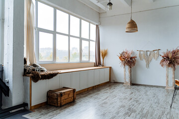 Interior shot. Bright room of the studio. A large panoramic window with a wide window sill, on which there is a vase with piles of wheat and a soft blanket.