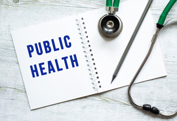PUBLIC HEALTH is written in a notebook on a wooden table next to stethoscope.
