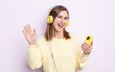 young pretty woman smiling happily, waving hand, welcoming and greeting you. headphones and phone concept © kues1