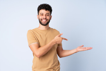 Young Moroccan man isolated on blue background holding copyspace imaginary on the palm to insert an...
