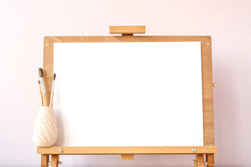 Simple mockup with blank white paper for painting and brushes on a wooden easel on table at home studio