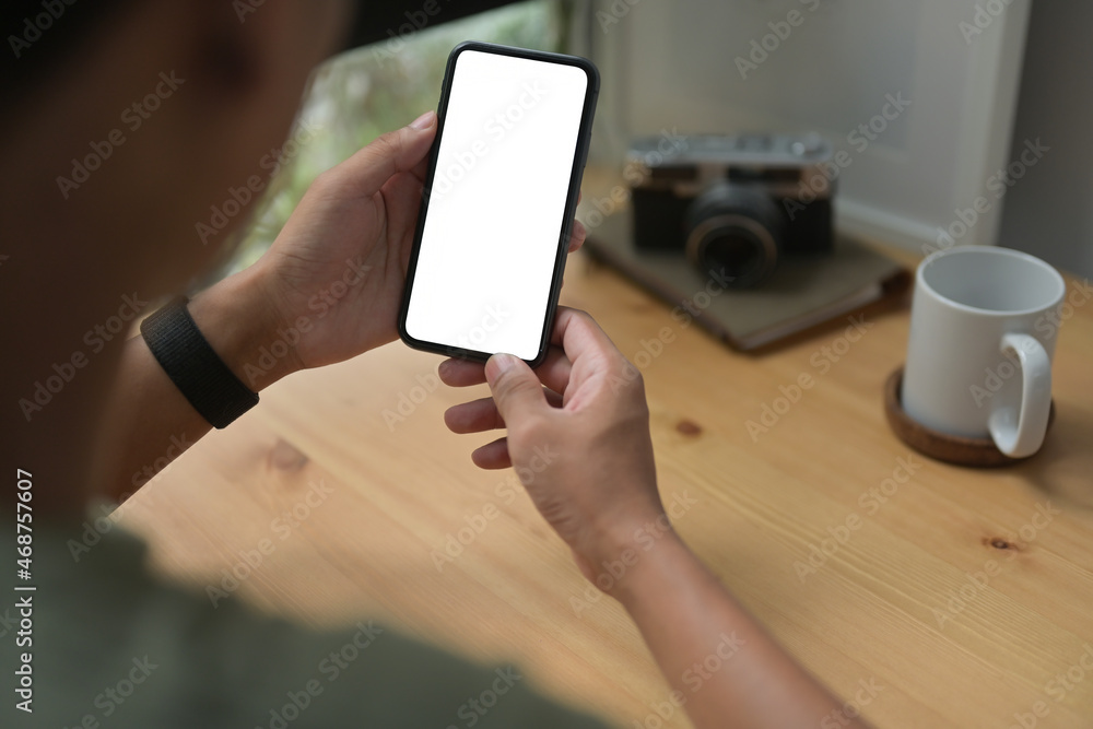 Poster behind view of a man using a white blank screen smartphone at the wooden working desk surrounded by 
