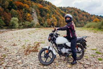 Fototapeta na wymiar Woman biker travel by motorbike in fall. Motorcyclist enjoys autumn landscape in mountains having rest by forest