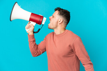 Young caucasian handsome man isolated on blue background shouting through a megaphone to announce something in lateral position