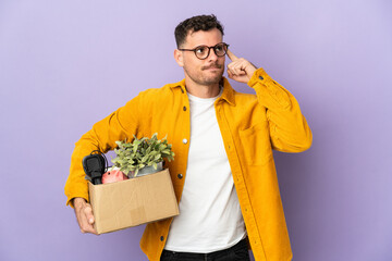 Young caucasian man making a move while picking up a box full of things isolated on purple...