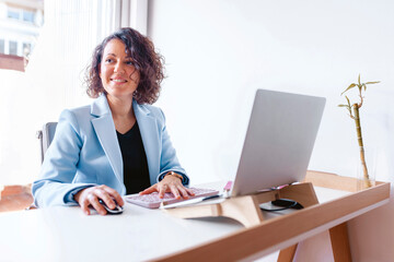 Cheerful woman working on her laptop at the office. Entrepreneur wearing blue suit using technology at work. Business woman, boss, manager, coworker, professional. Business concept.