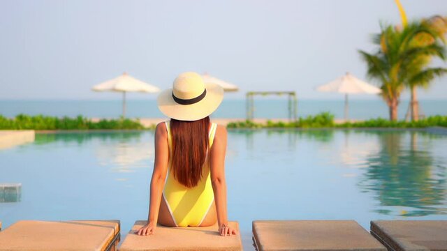 Woman sitting on the edge of the swimming pool at an exotic hotel in Florida wearing yellow monokini and floppy hat looking at the beach, back view slow-motion handheld