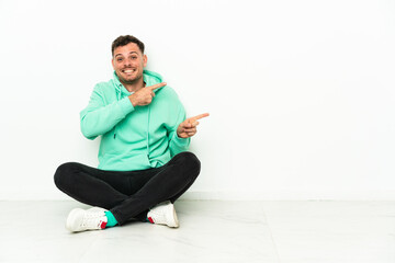 Young handsome caucasian man sitting on the floor surprised and pointing side
