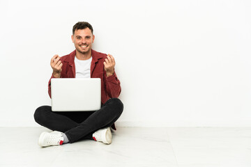 Young handsome caucasian man sit-in on the floor with laptop making money gesture