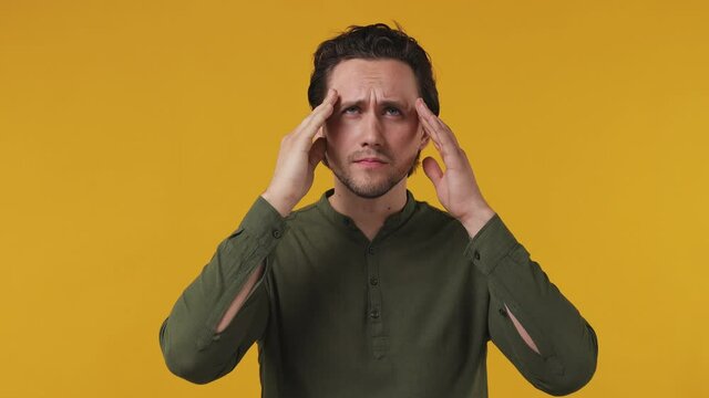 Sick ill sad tired jaded young brunet bearded man 20s wears green shirt put hands on head rub temples having headache suffering from migraine feel bad seedy isolated on pastel plain yellow background
