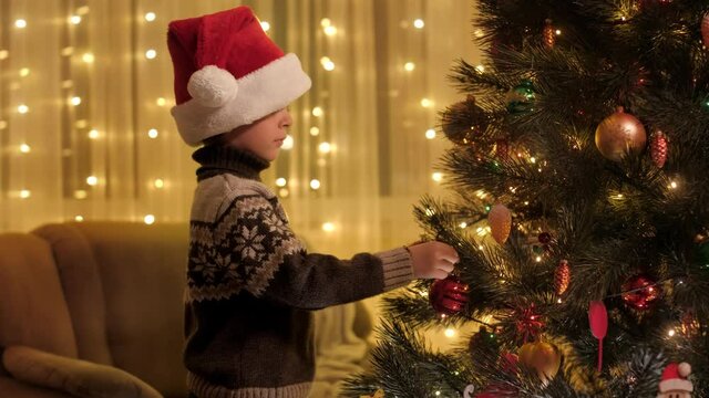 Portrait of little boy hanging red bable on glowing colorful Christmas tree in living room. Pure emotions of families and children celebrating winter holidays.