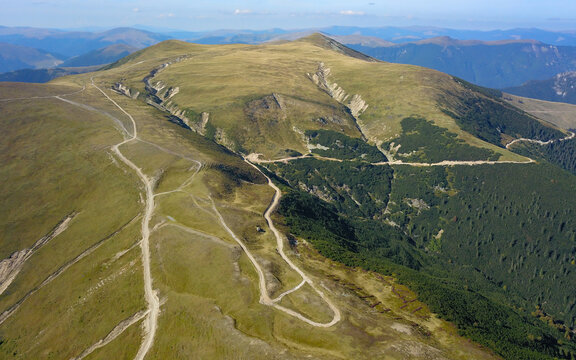 Aerial drone view of a gravel road winding at a high altitude to Nedeia Peak and the alpine pastures and forests surounding it. Capatanii Mountains, Romania.