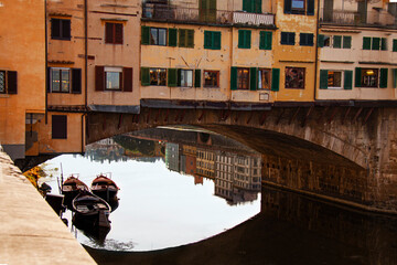 View of the Ponte Vecchio - Old Bridge - in Florence (Firenze) - by the river Arno with blue sky and reflections and boats in the water. Famous landmark in Tuscany, Italia. - obrazy, fototapety, plakaty