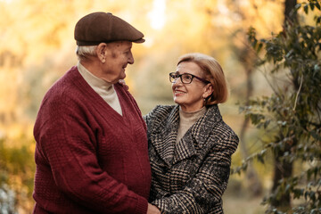 Fototapeta premium Elderly man looking at his elegant retired woman with love, warming her hands and enjoying her beauty, senior married couple dancing in autumn park, celebrating anniversary, affection
