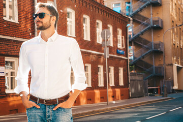 Fototapeta premium Portrait of handsome confident stylish hipster lambersexual model.Modern man dressed in white shirt. Fashion male posing on the street background in sunglasses. Outdoors at sunset