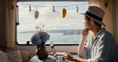 Young man wearing hat in campervan typing on laptop with beatiful ocean view