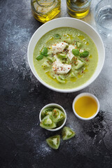 Green tomato gazpacho soup served with mozzarella in a white bowl, flatlay on a dark-grey stone background, vertical shot