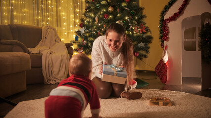 Obraz premium Little baby boy in Santa Claus costume crawling to his mother sitting under Christmas tree. Families and children celebrating winter holidays.