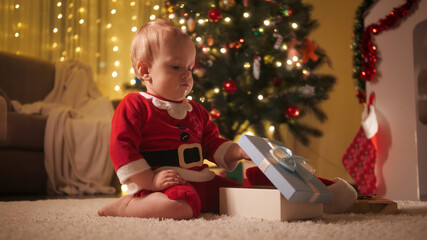 Fototapeta na wymiar Little baby boy in Santa Claus costume opens Christmas gift box. Families and children celebrating winter holidays.
